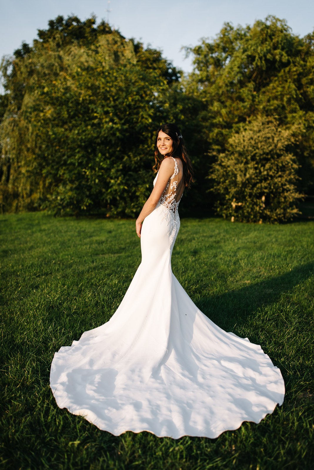 Bride looking back over her shoulder, her dress train fanned out on the grass in the evening sun