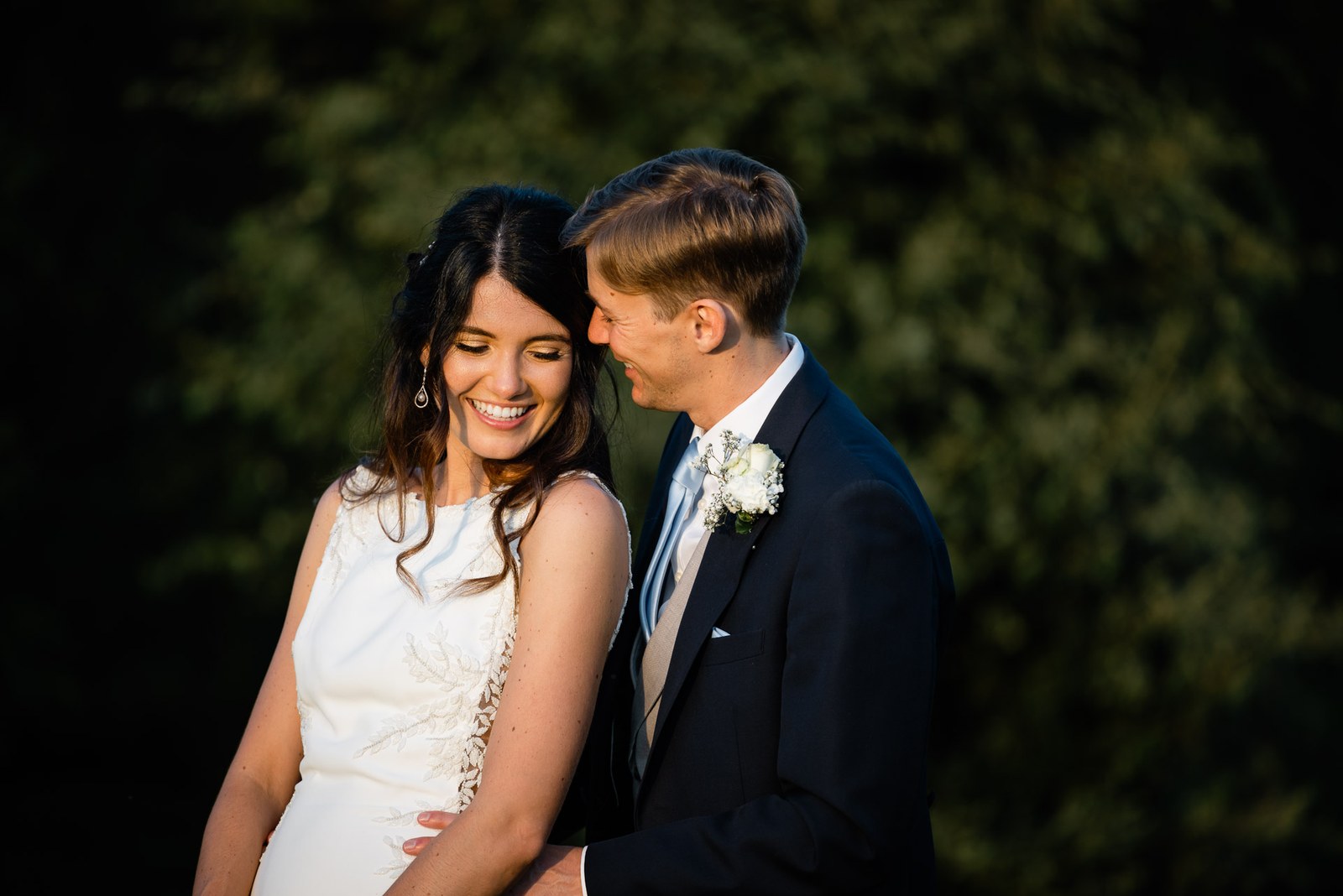 The couple laughing together against dark summer foliage at dusk