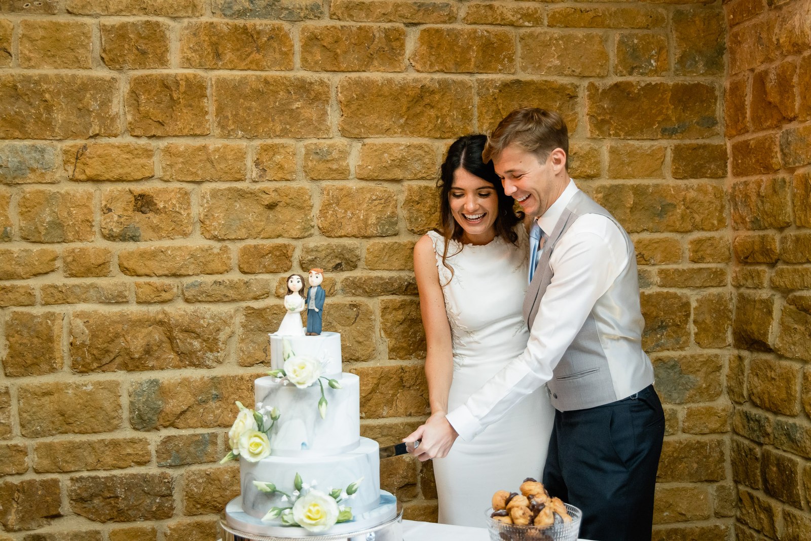 The couple cutting their three-tier cake against the honey-stone wall at Dodford Manor