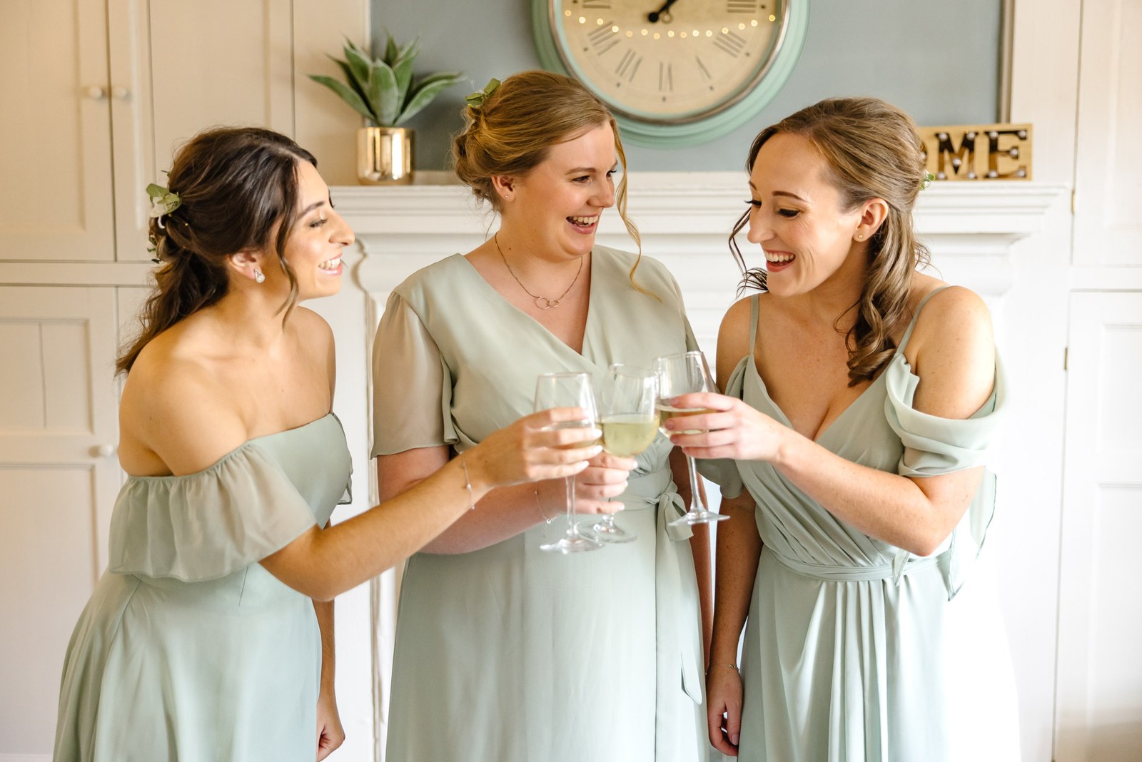 Bridesmaids in sage-green dresses toasting with champagne beside a mantelpiece clock