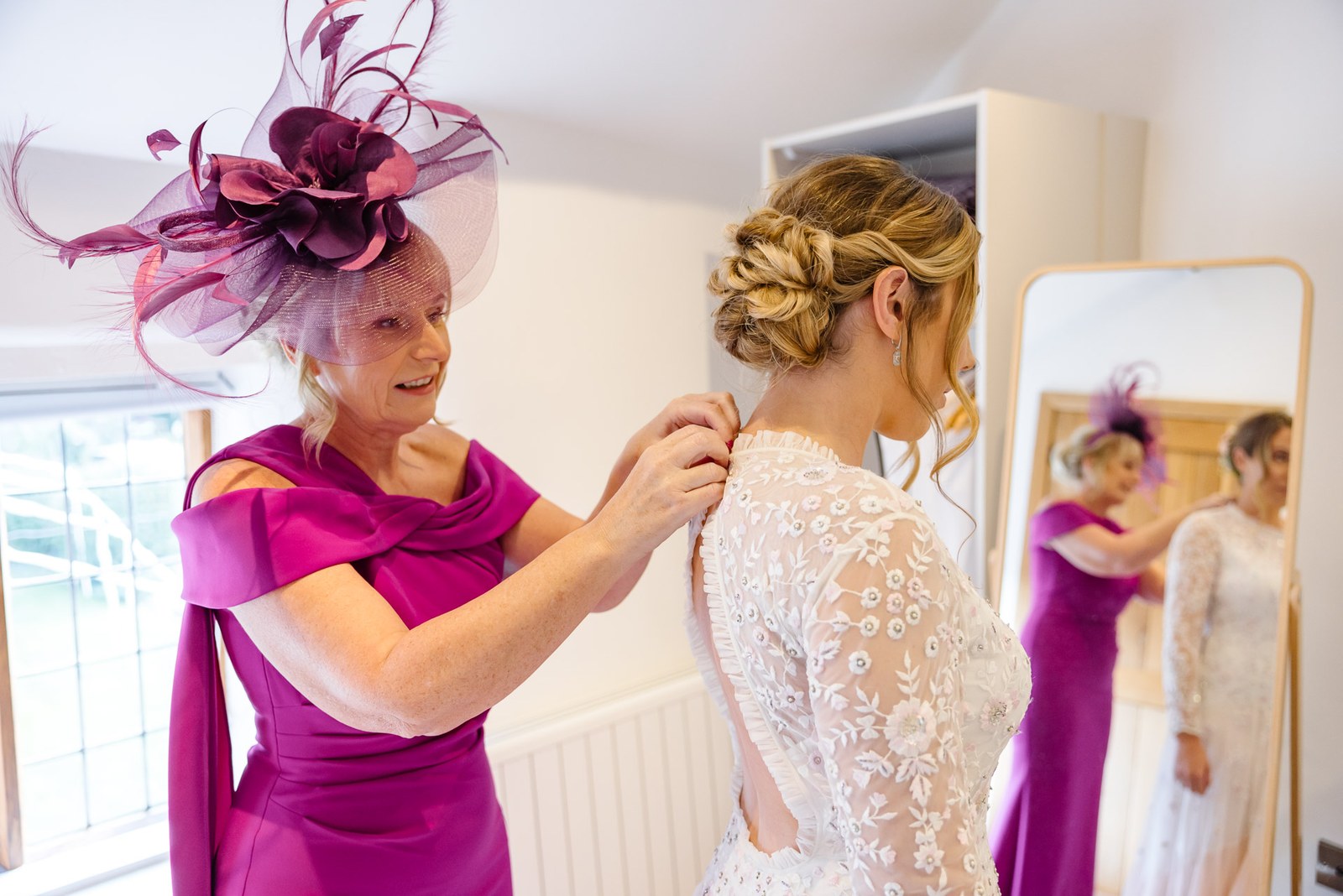 Mother of the bride in magenta fastening the back of the bride's beaded lace gown