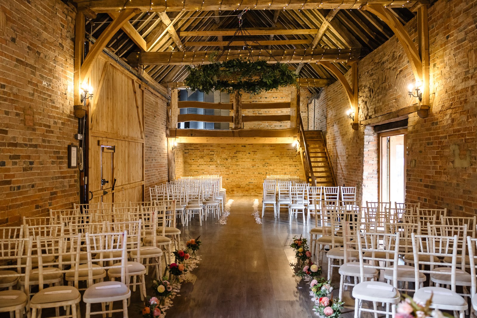 The exposed-brick ceremony barn at Dovecote Events, chairs set and the aisle lined with flowers