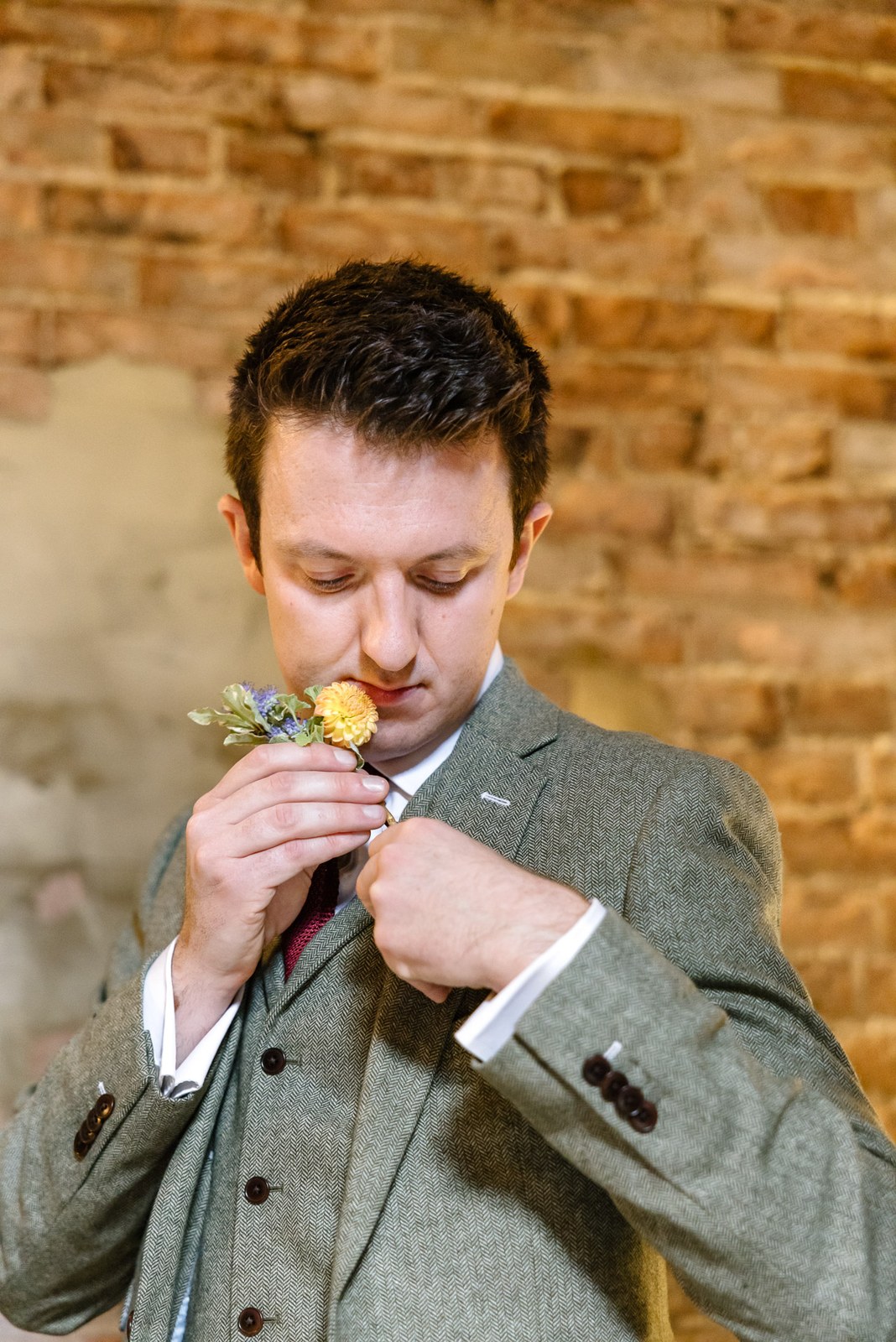 Groom in a green tweed suit breathing in the scent of his buttonhole against a brick wall