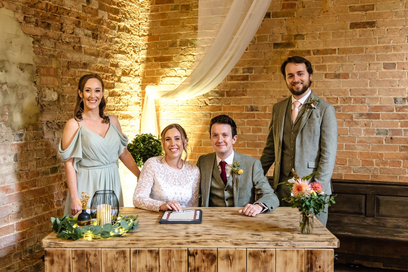 The newlyweds and their witnesses at a rustic register table against the draped brick wall