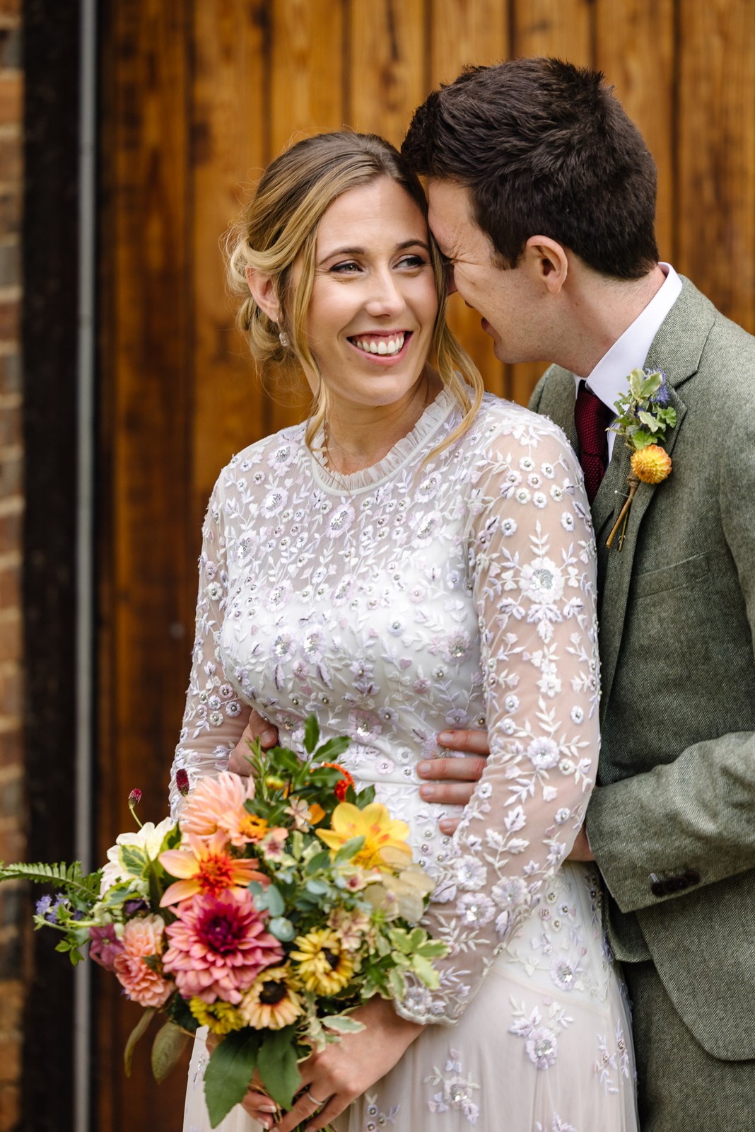 The couple close together by a timber barn door, the bride holding a bright autumn bouquet