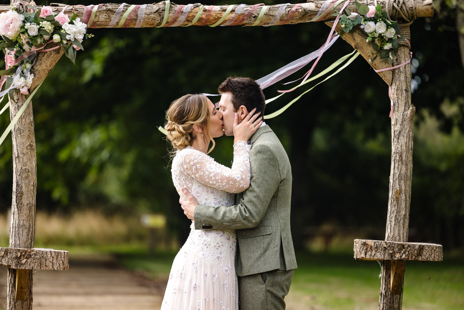 The couple kissing under a flower-and-ribbon wooden arch in the Dovecote Events grounds