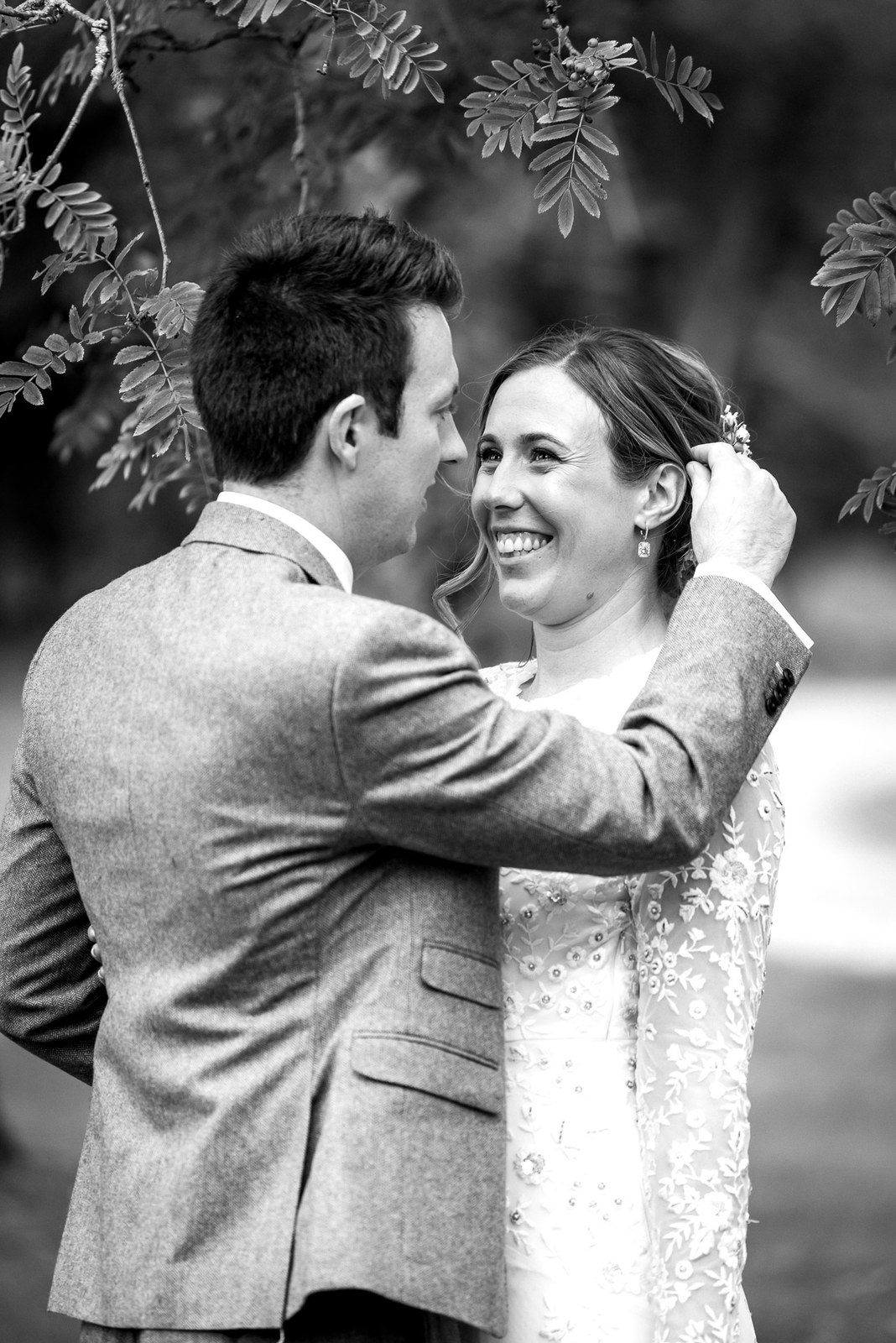 Black-and-white moment of the groom smoothing the laughing bride's hair beneath a tree