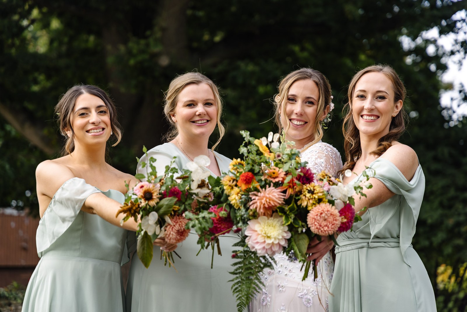 Bridesmaids in sage green gathered around the bride, all holding autumnal bouquets