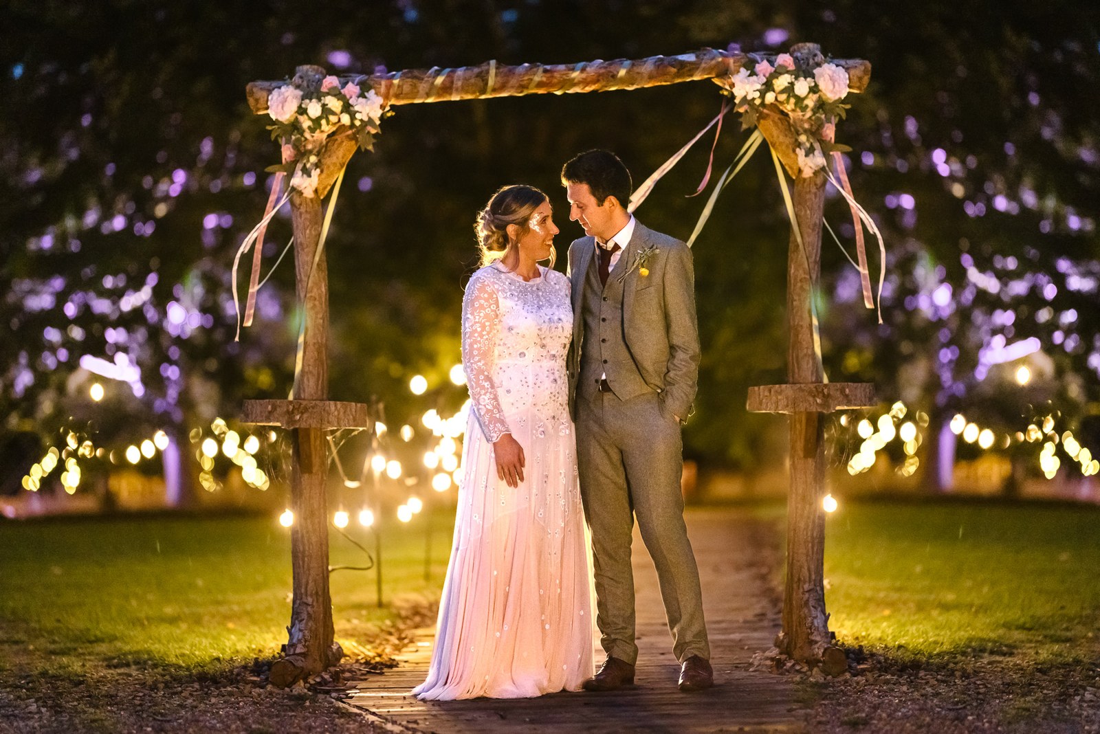 The couple under the lit wooden arch at night, fairy lights glowing behind at Dovecote Events