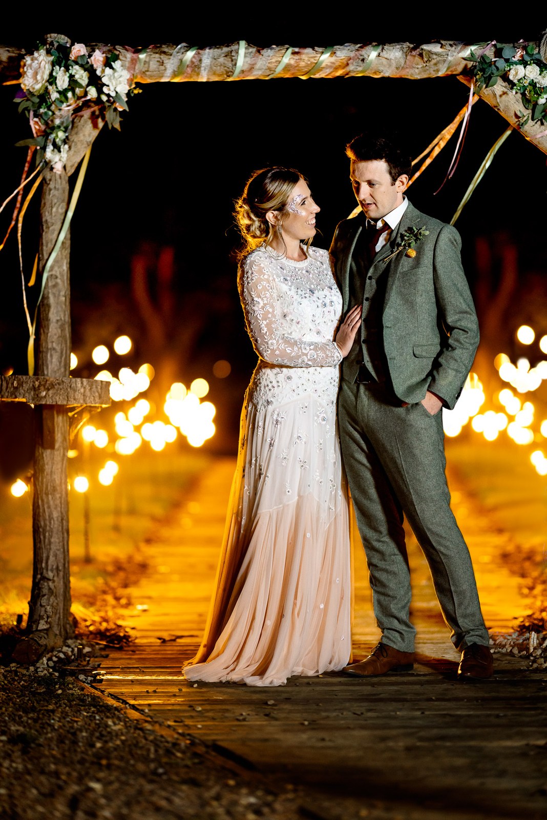 Evening portrait of the couple under the ribboned arch, fairy lights lining the Dovecote Events boardwalk