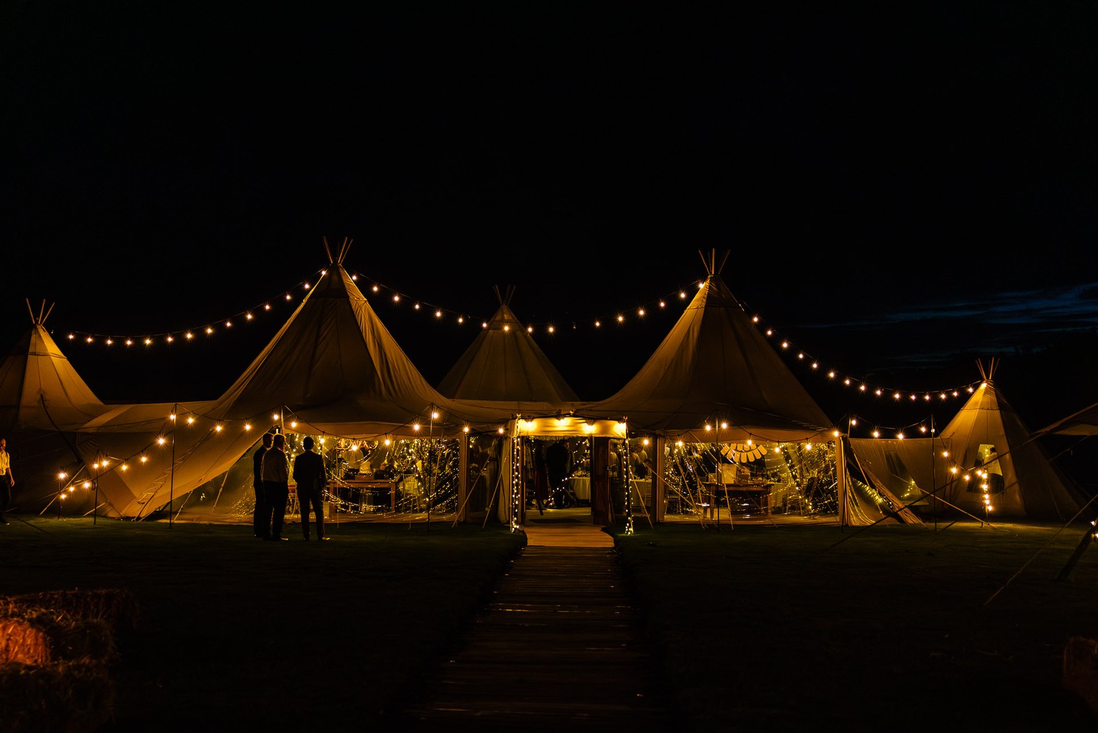 The illuminated tipis at Dovecote Events glowing at night, festoon lights strung between the peaks