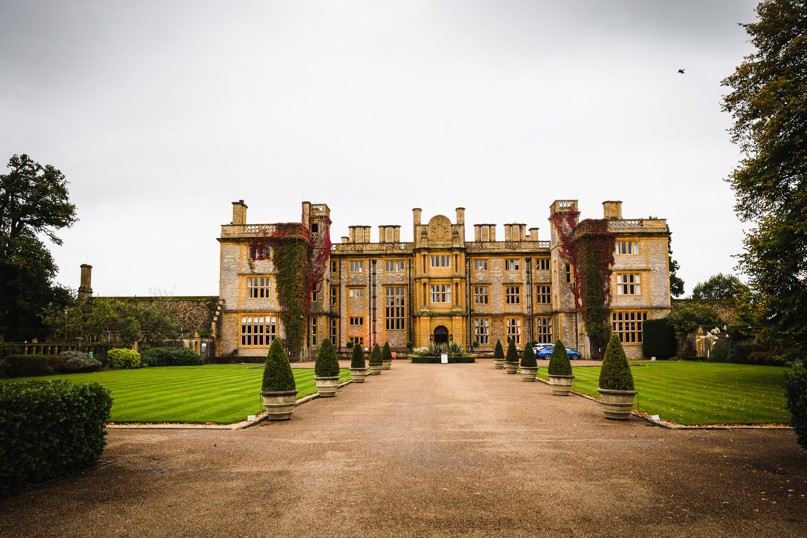 The Jacobean stone facade of Eynsham Hall, formal lawns and topiary lining the gravel drive