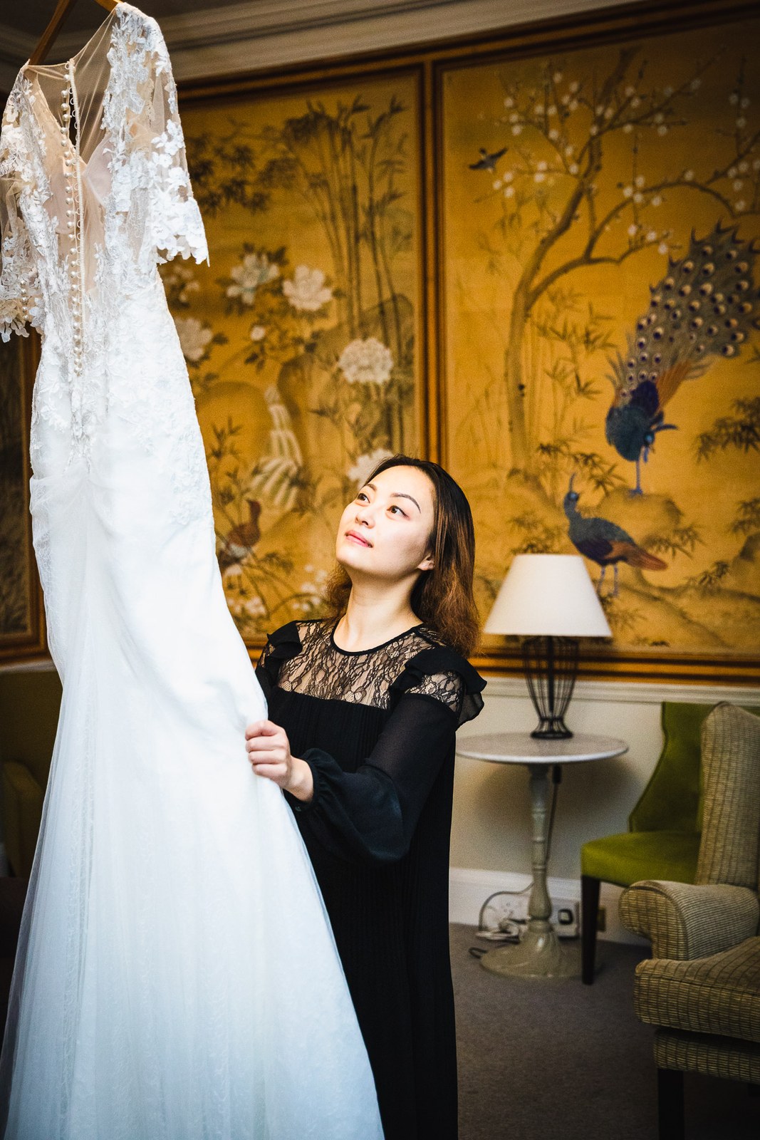 A woman holding up the lace gown against gold Chinoiserie wallpaper in a suite at Eynsham Hall