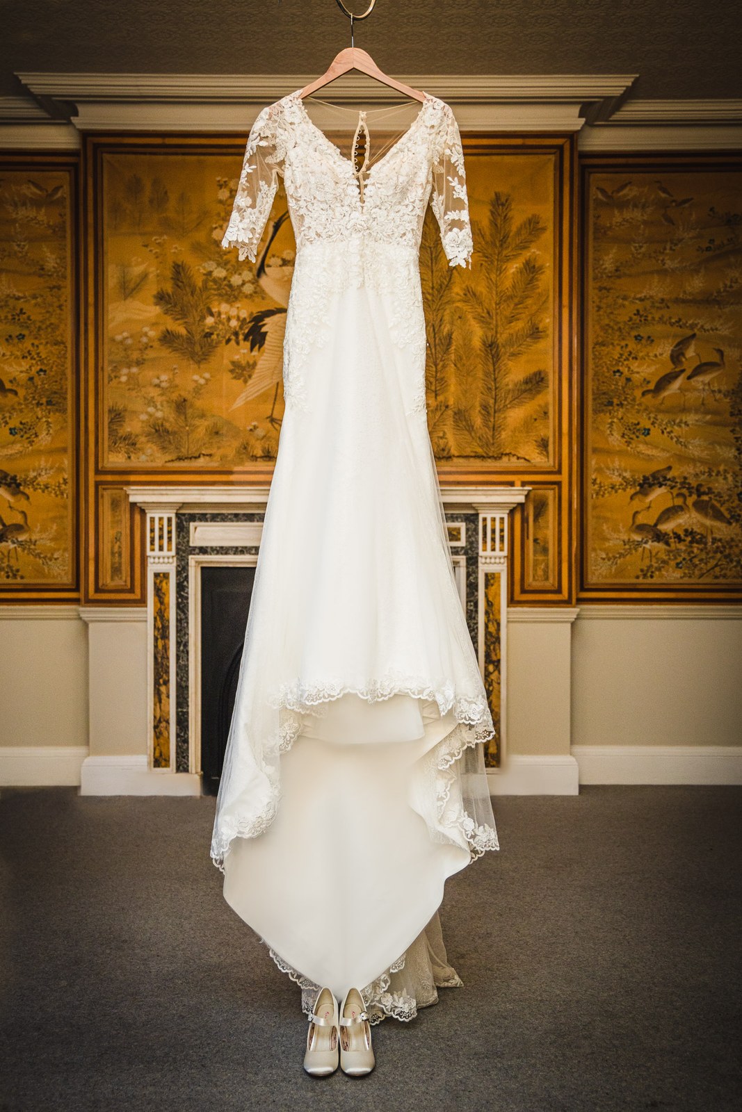 The lace wedding gown hanging before gilded peacock wallpaper and a marble fireplace at Eynsham Hall