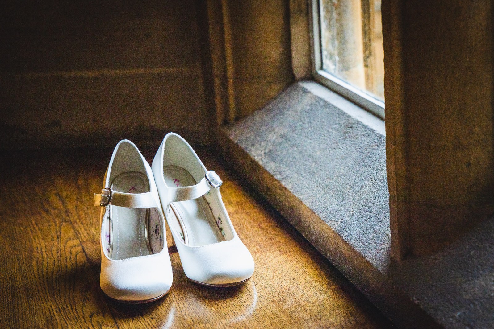 A pair of white Mary-Jane bridal shoes on a sunlit wooden floor beside a stone window
