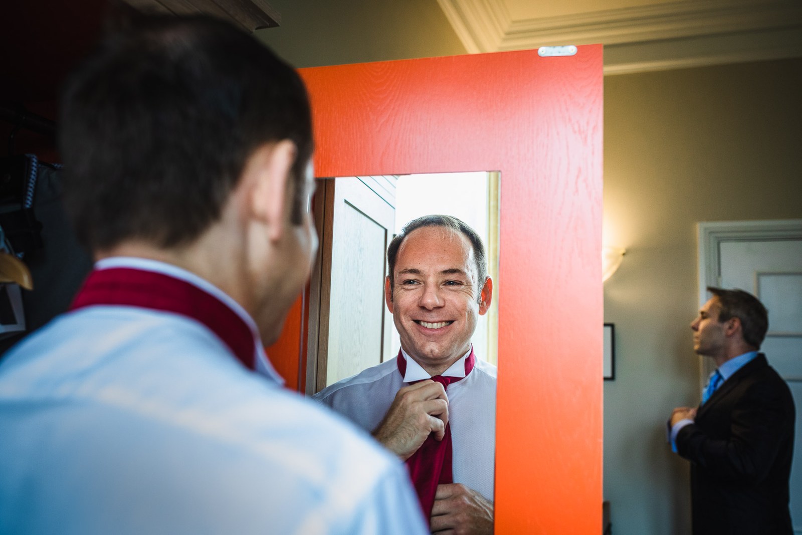 Groom adjusting his tie in a mirror while a groomsman waits behind