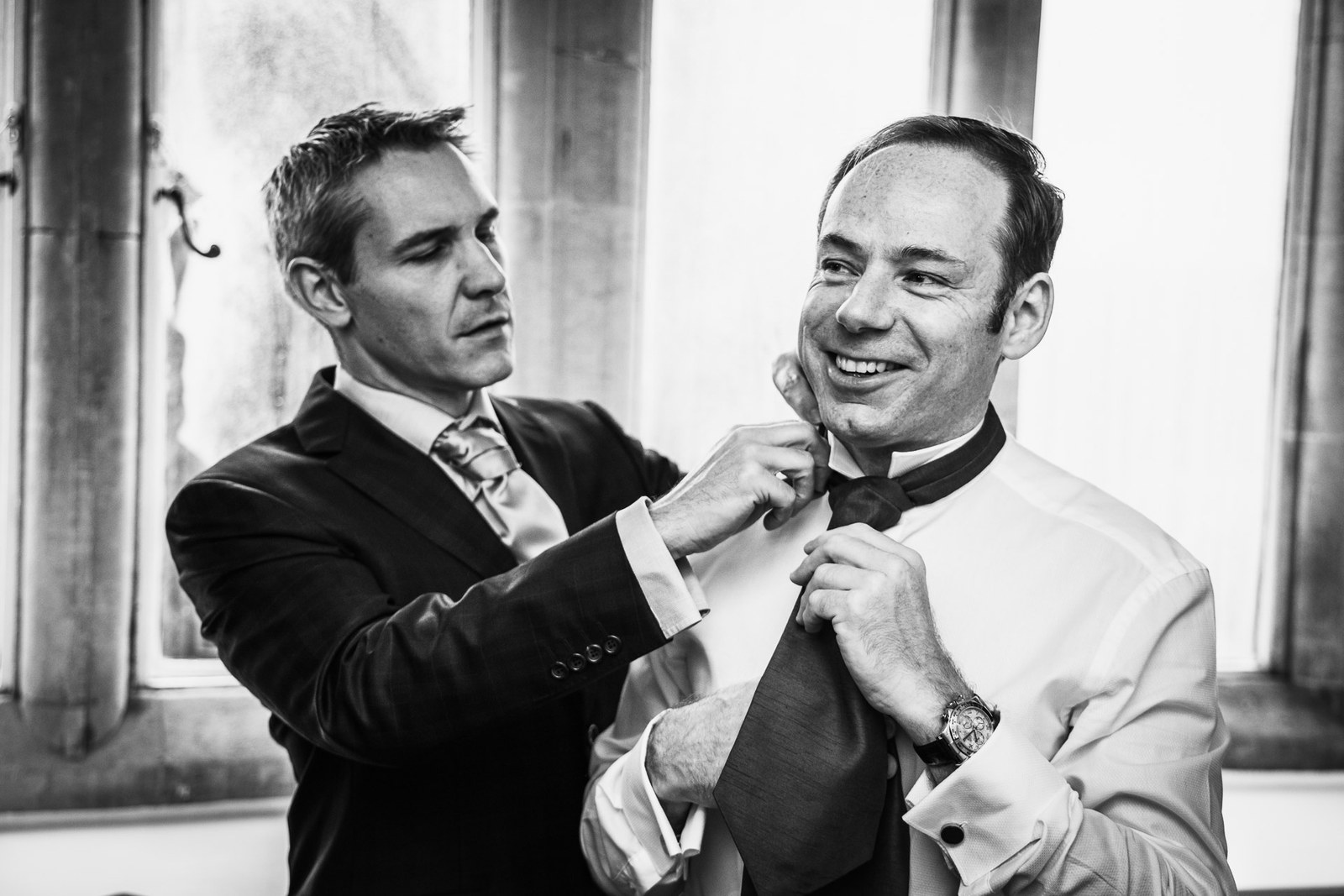 Black-and-white of a groomsman straightening the groom's collar and tie by a window