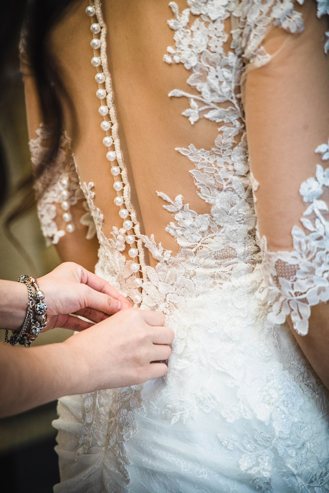 Hands doing up the long row of pearl buttons down the back of the bride's lace gown