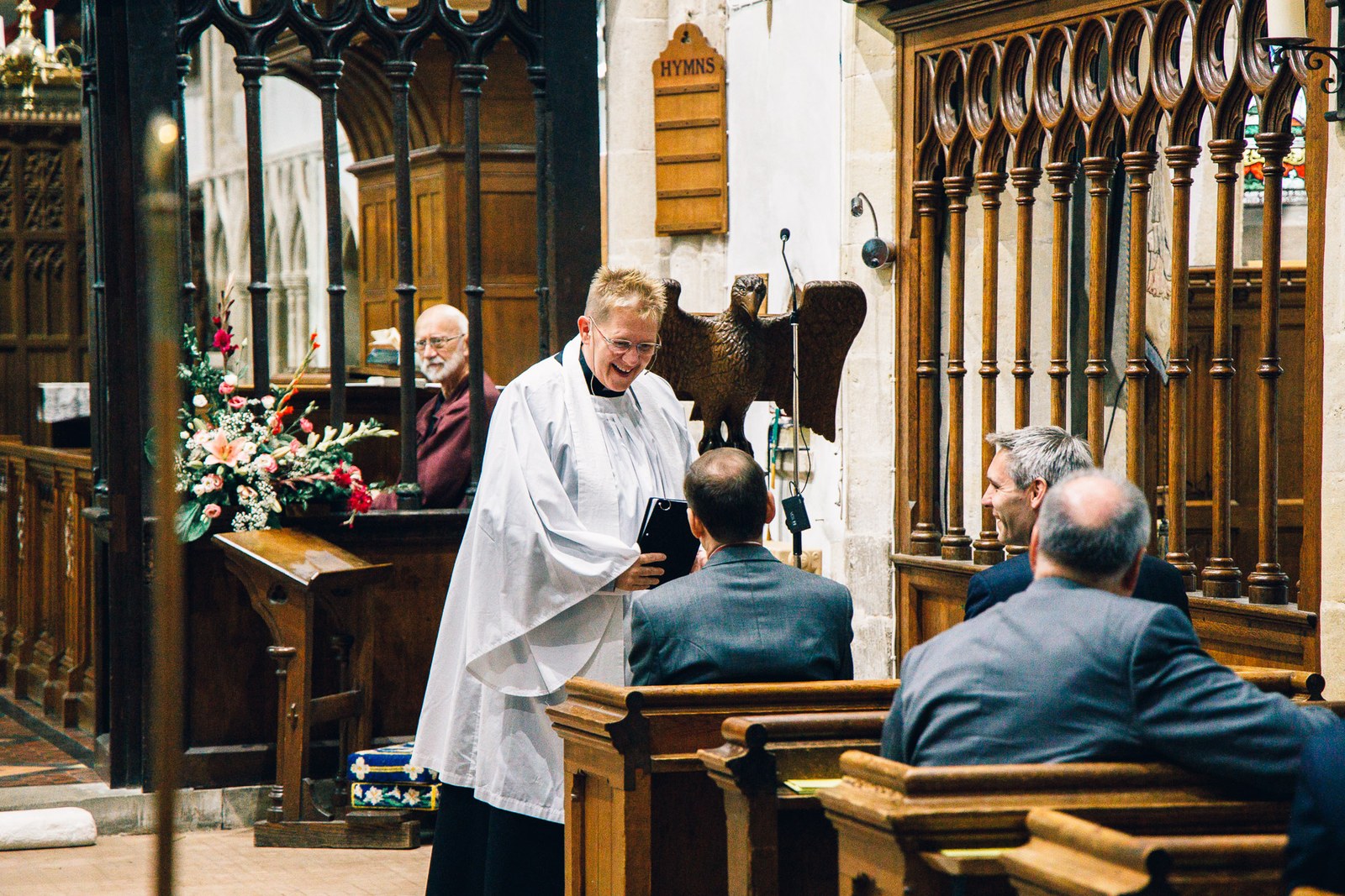 The vicar in white robes laughing beside the carved eagle lectern before the service