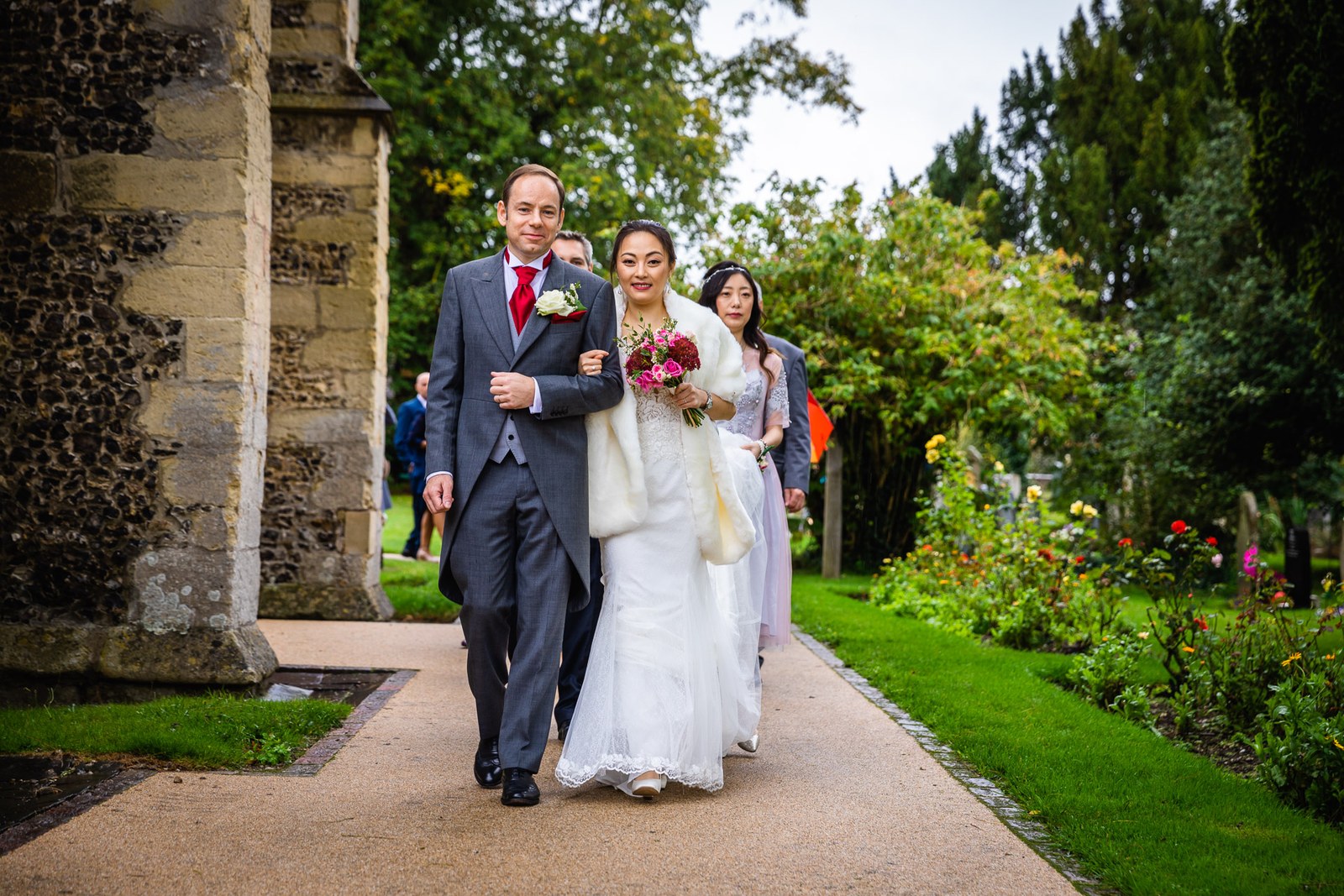 Bride and groom arm in arm along the rose-lined church path, a bridesmaid behind