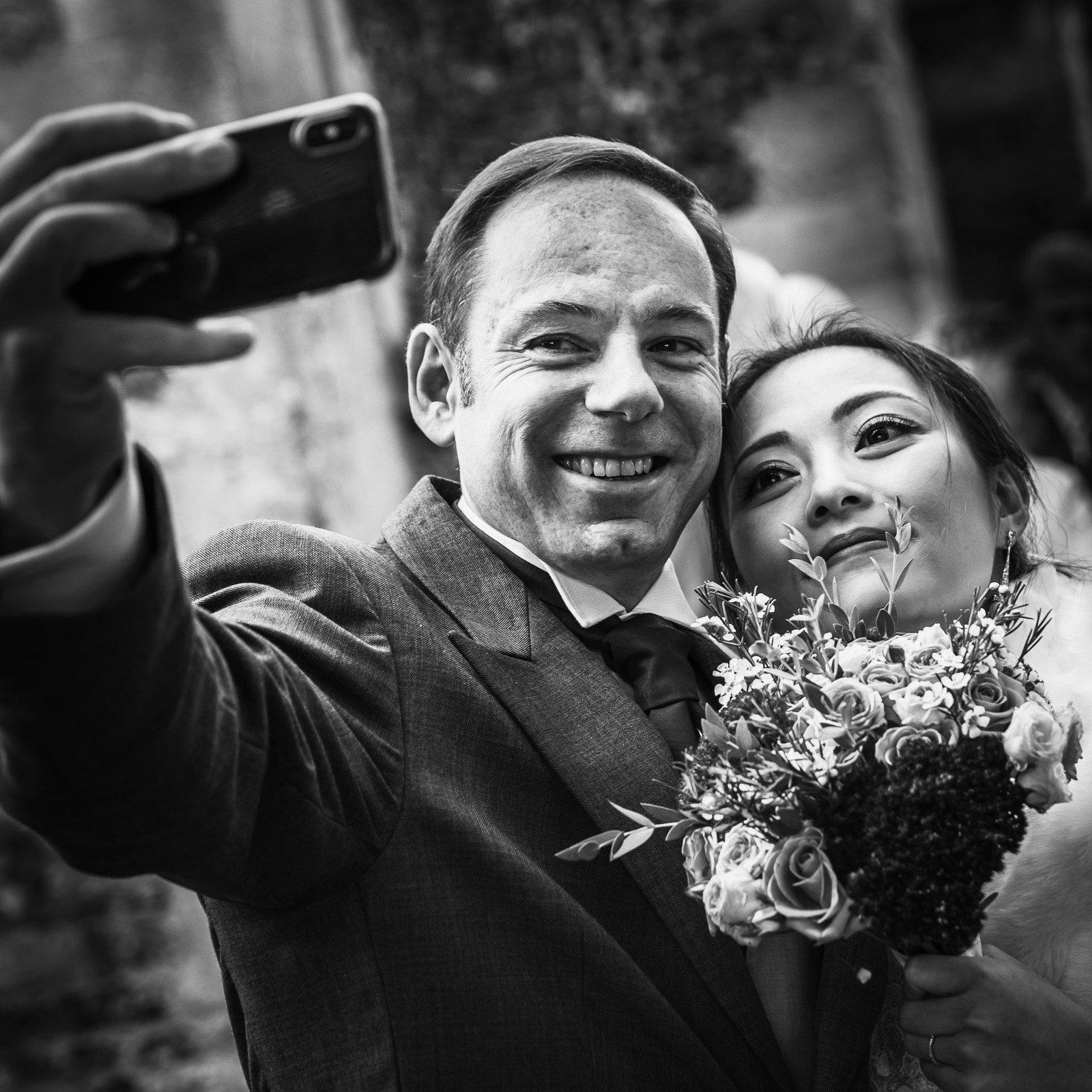 Black-and-white of the groom taking a selfie with his bride, bouquet in hand