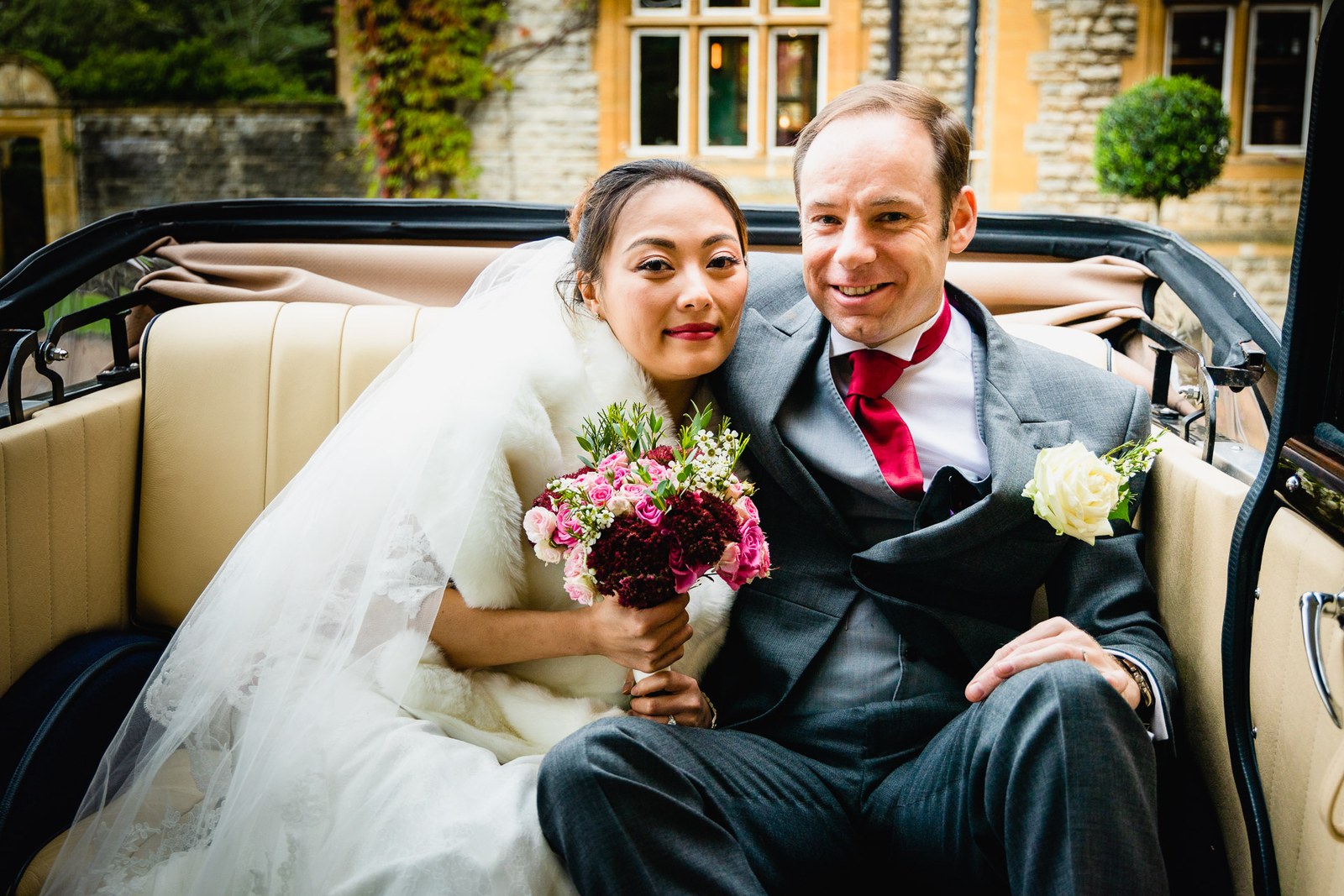 Bride and groom in the open vintage car before the honey-stone facade of Eynsham Hall