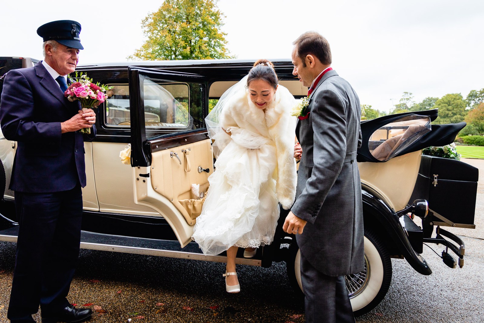 The chauffeur helping the bride from the car as the groom waits