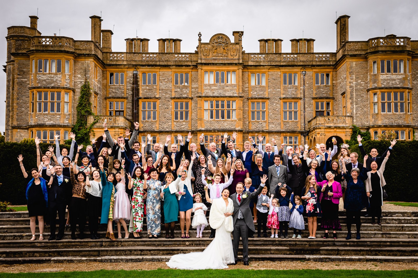 The whole wedding party cheering on the steps before the Jacobean front of Eynsham Hall