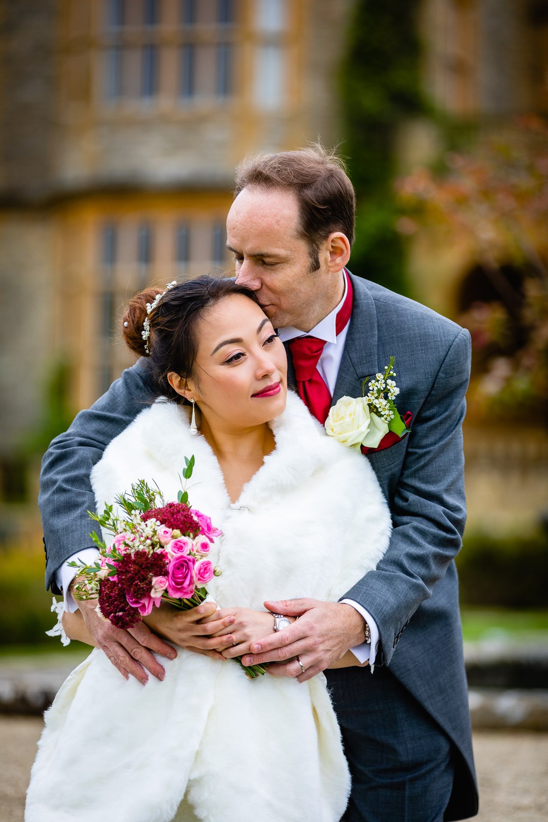 Groom kissing the bride's head as she leans into him, Eynsham Hall rising behind