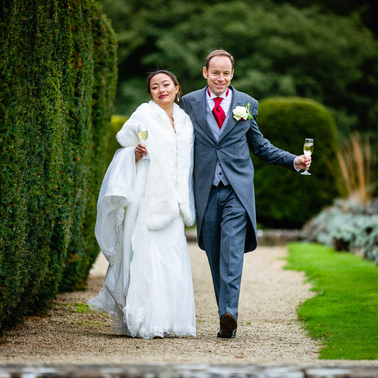 The couple walking with champagne through the clipped topiary gardens at Eynsham Hall
