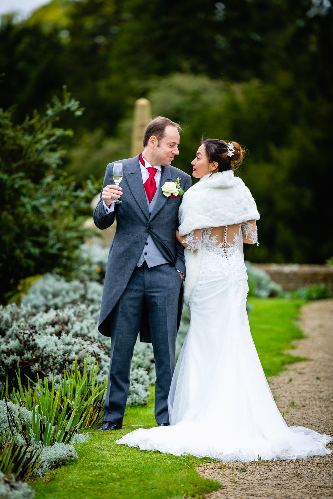 The couple pausing with champagne on a garden path, the bride in a fur wrap