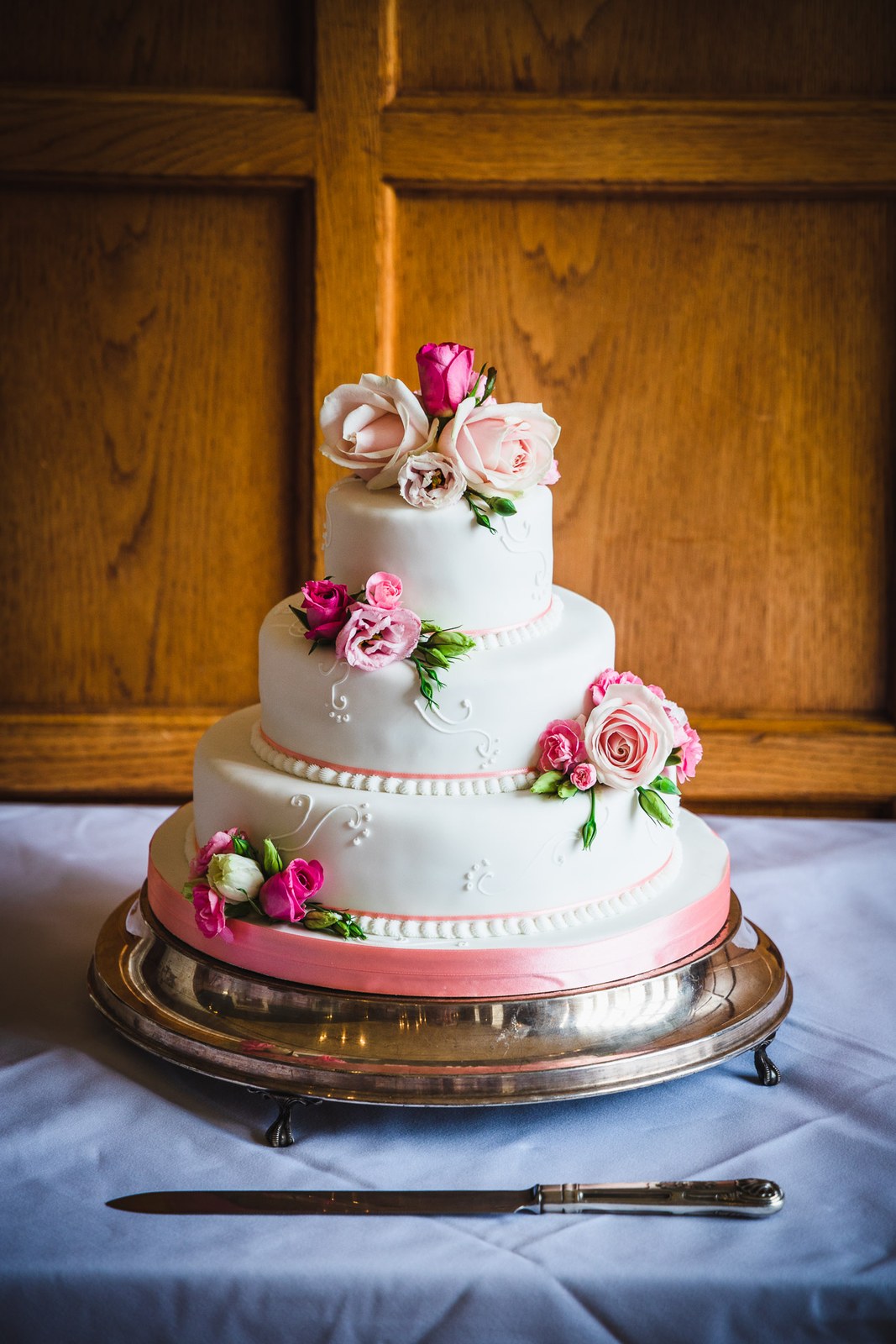 The three-tier cake with pink ribbon and roses on a silver stand at Eynsham Hall