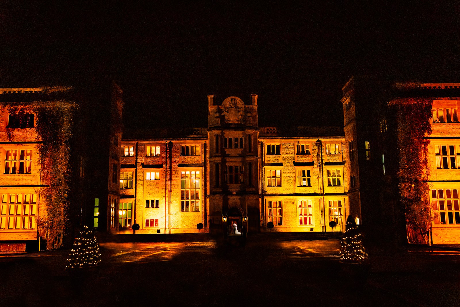 Eynsham Hall floodlit amber at night, the couple tiny before its grand facade