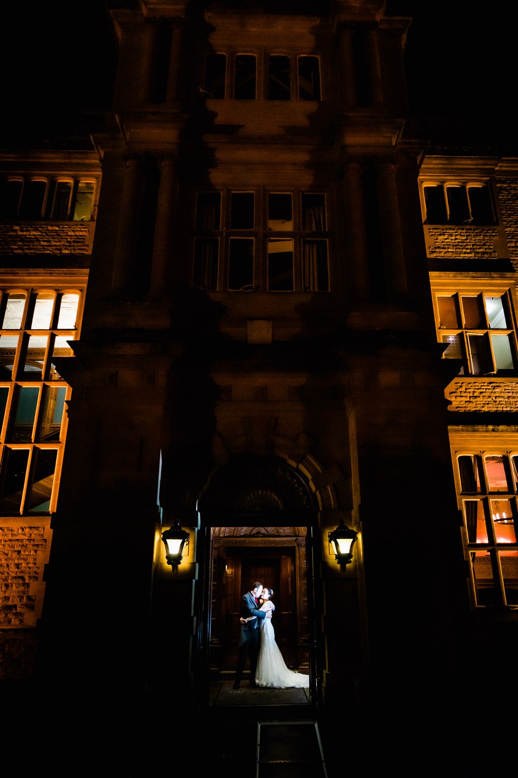 The couple embracing in the lit archway entrance of Eynsham Hall at night