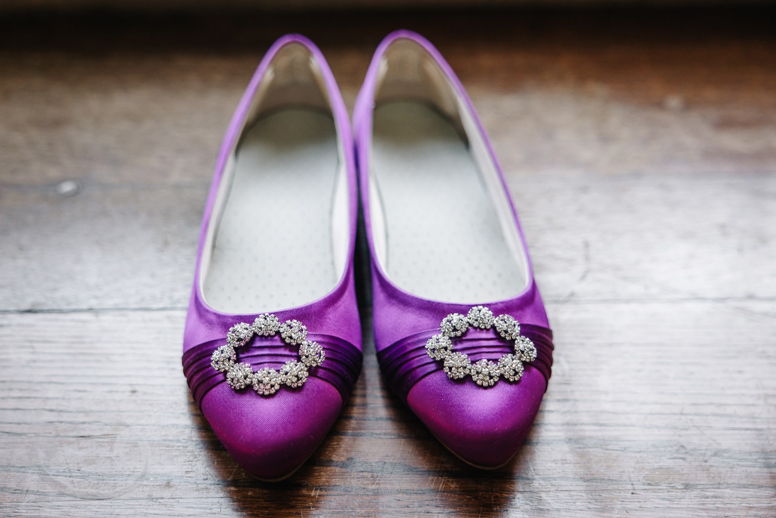 A pair of purple satin bridal shoes with diamante buckles on a wooden floor