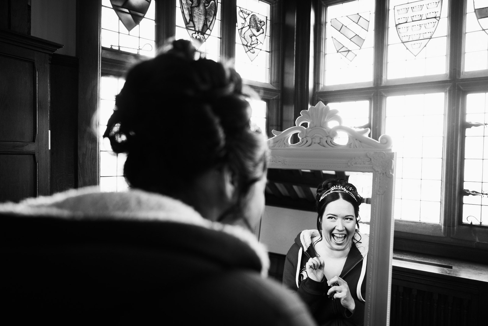 Black-and-white of the bride laughing at her reflection in a mirror by stained-glass windows
