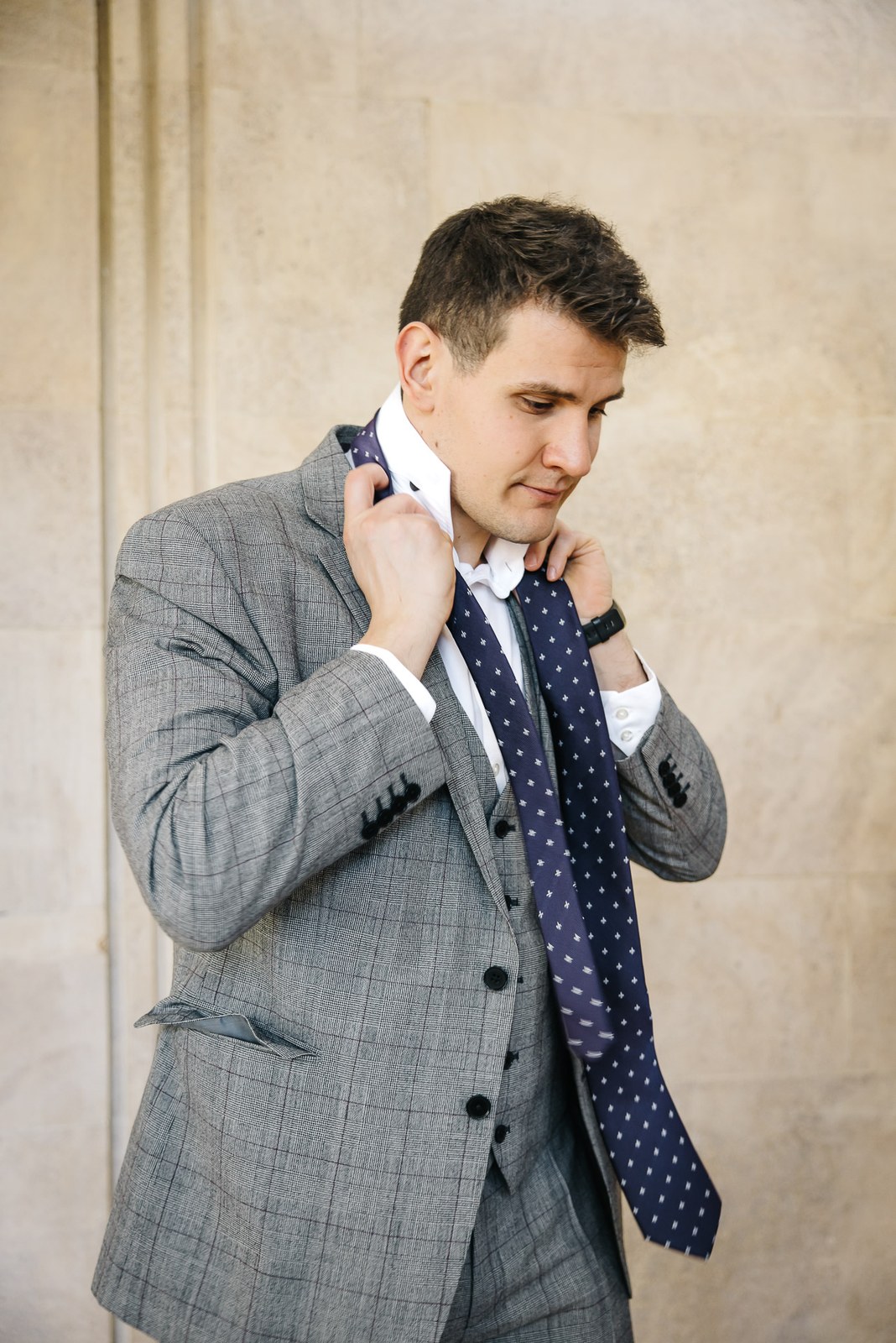 Groom knotting his polka-dot tie against a pale stone wall