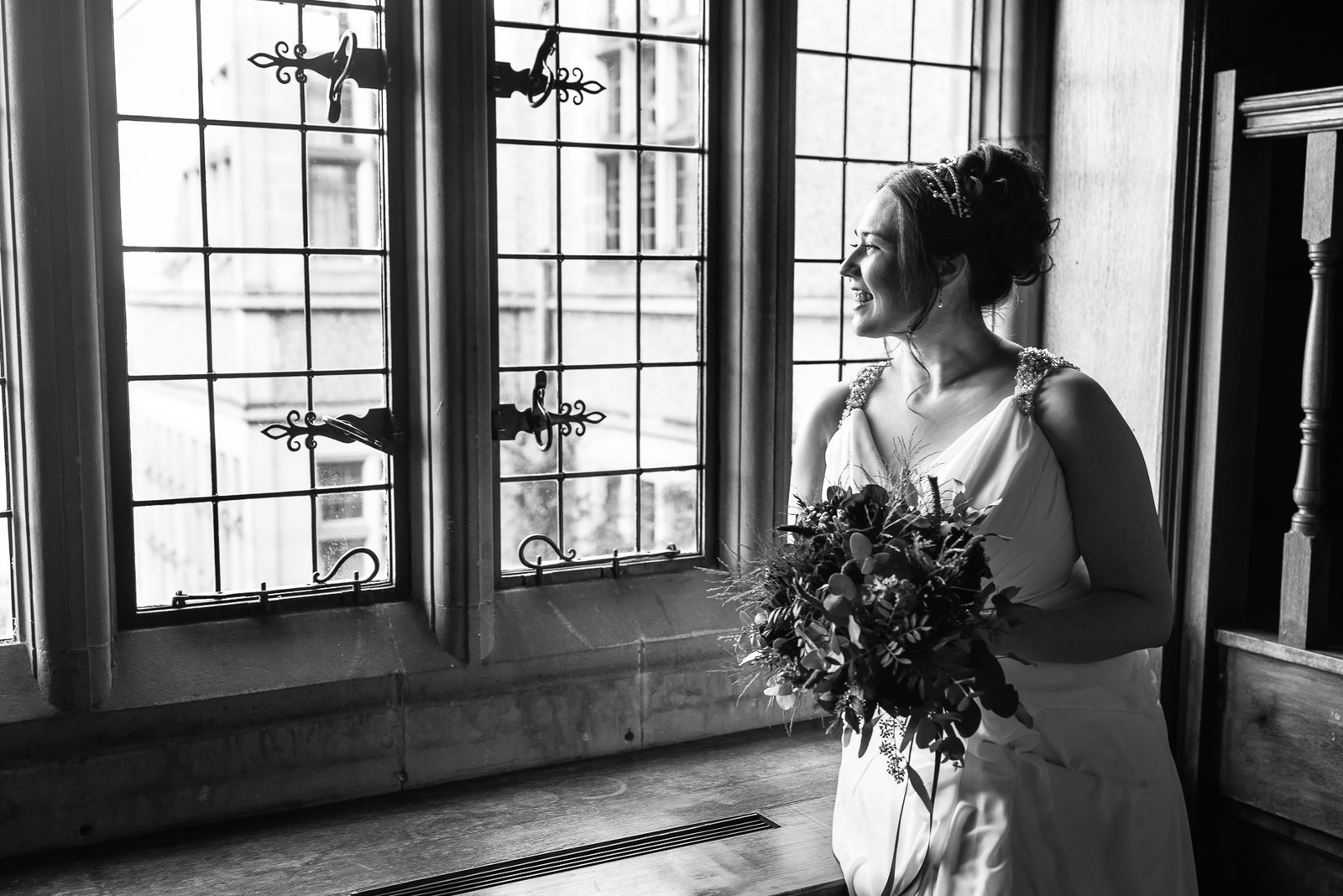 Black-and-white of the bride in profile at a tall leaded window, bouquet in hand