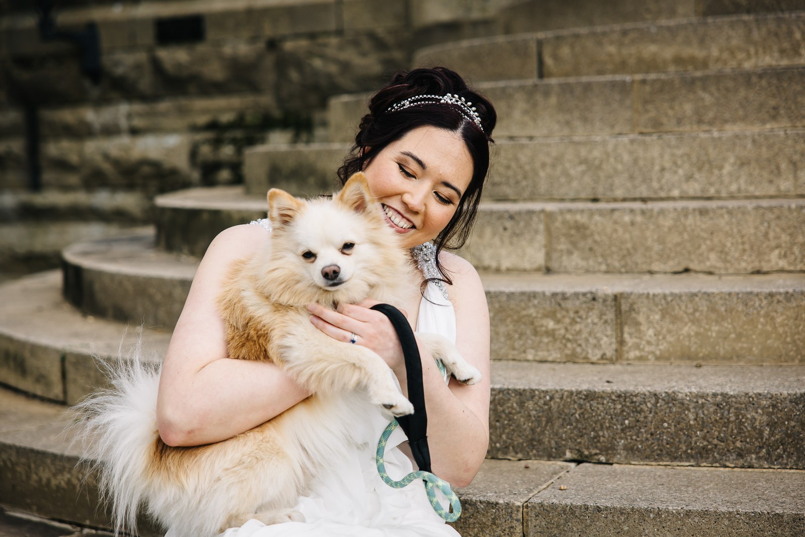 Bride sitting on stone steps cuddling a small fluffy dog