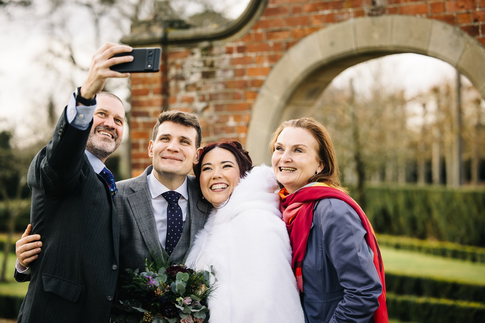 A guest taking a group selfie with the bride and groom by a brick garden arch