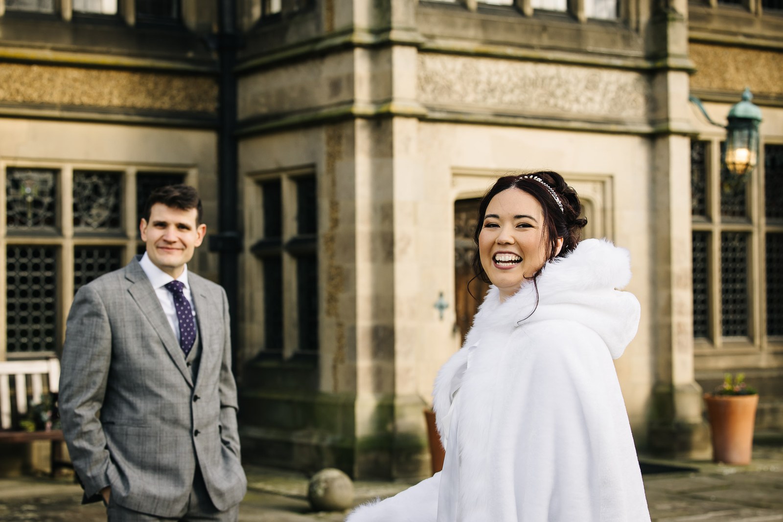 Bride turning to laugh beside the stone facade of Fanhams Hall, wrapped in a white fur cape