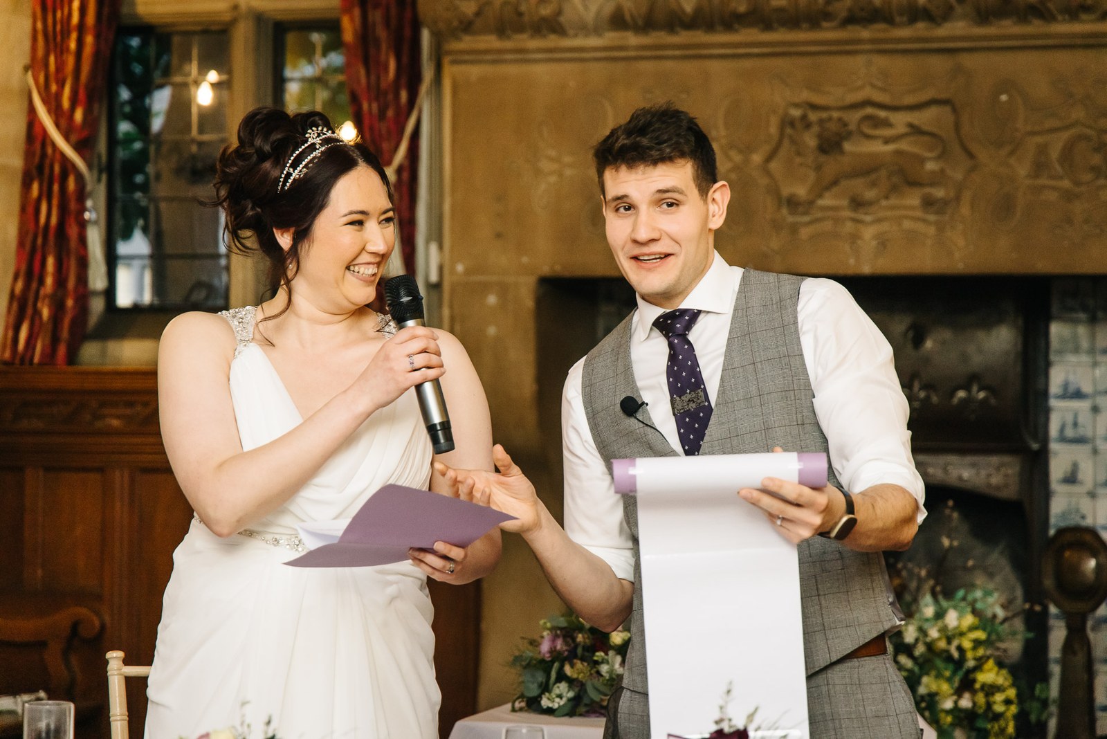 Bride and groom sharing a microphone for their speech before the carved fireplace at Fanhams Hall