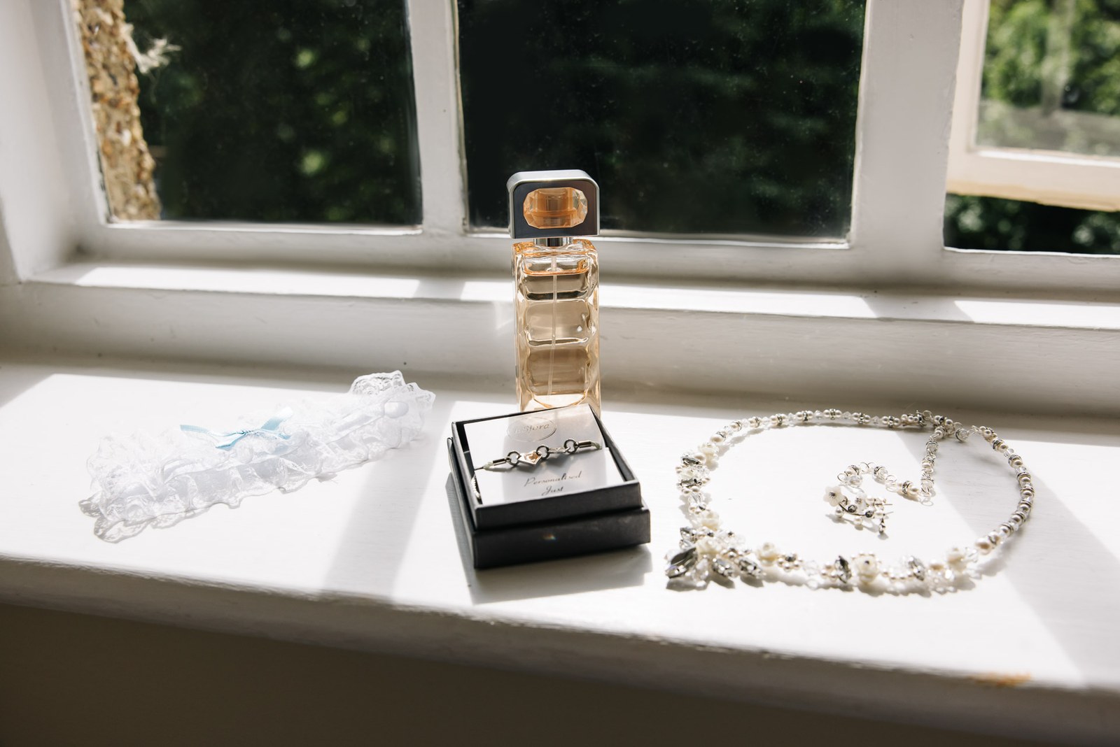 Bridal details on a windowsill - perfume, boxed earrings, a garter and a necklace laid in a heart