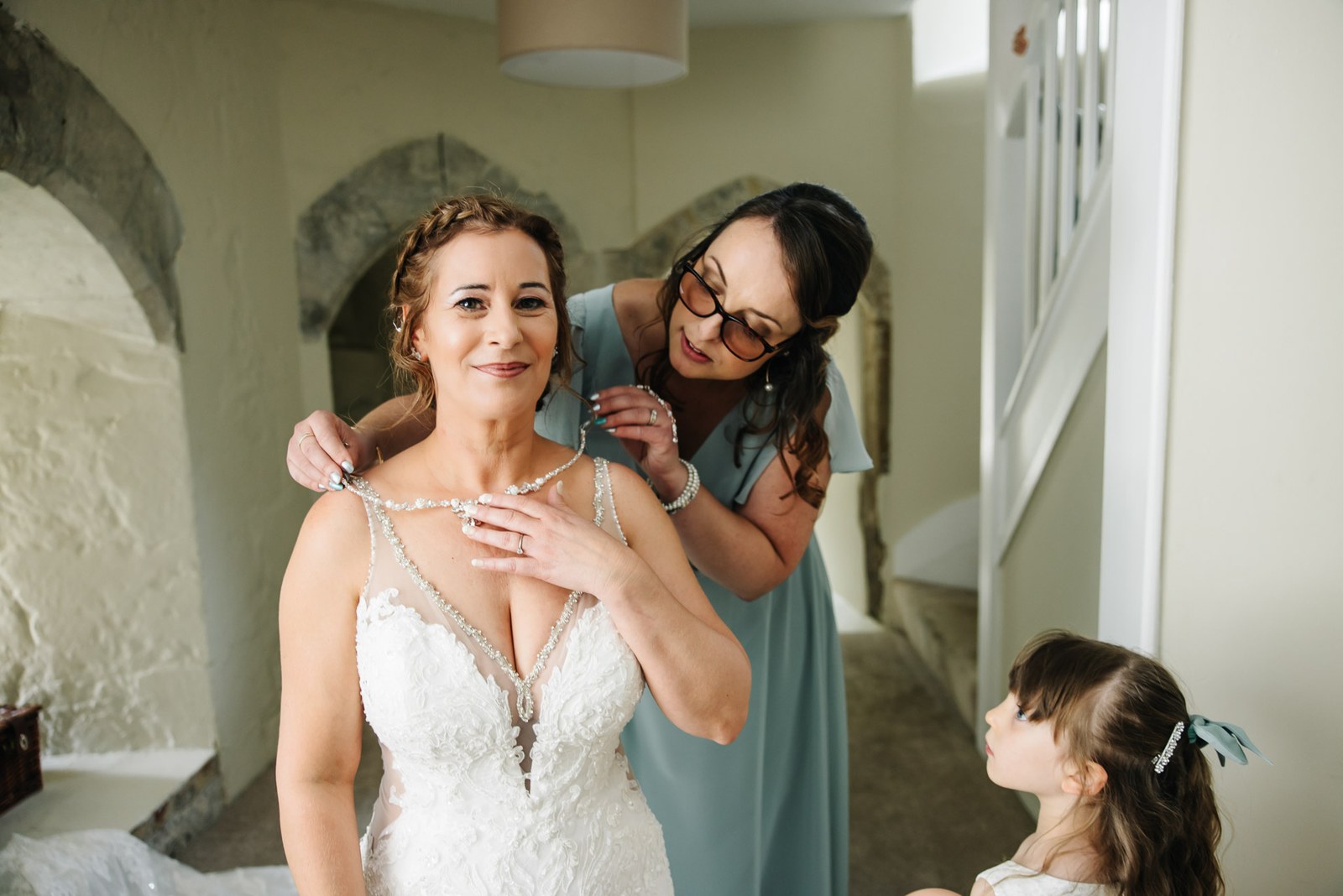 A bridesmaid fastening the bride's necklace as a flower girl looks up