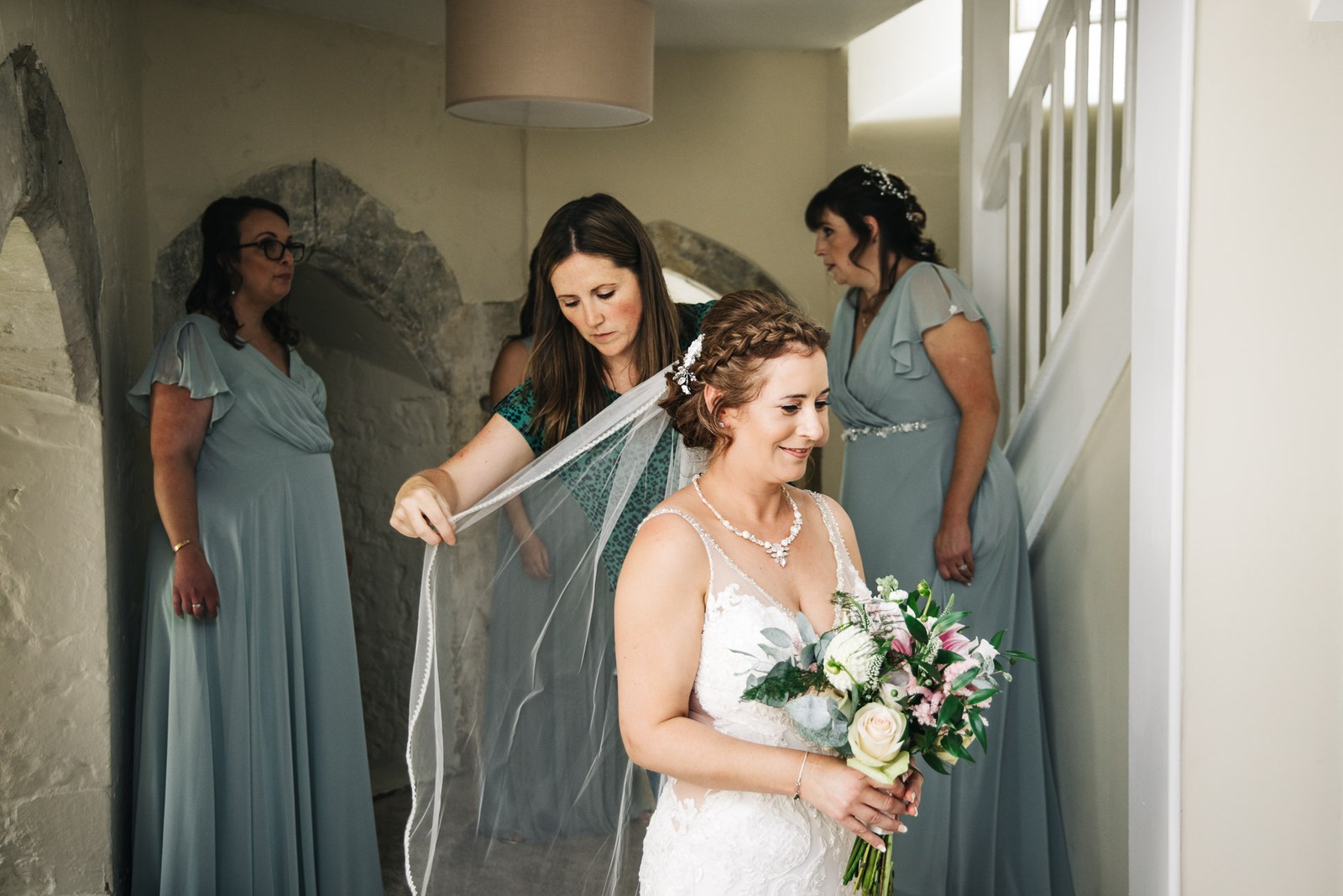 A bridesmaid arranging the bride's veil in a stone-arched room, bouquet in hand
