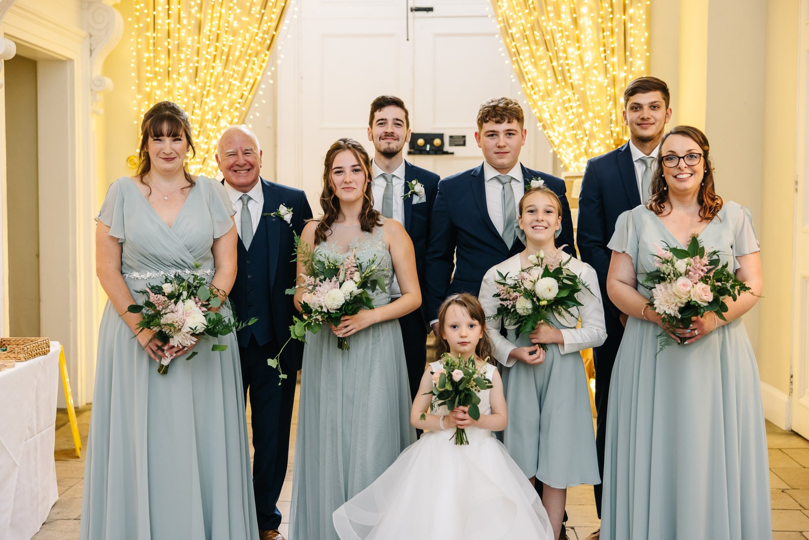The wedding party gathered before a fairy-light backdrop, bouquets in hand