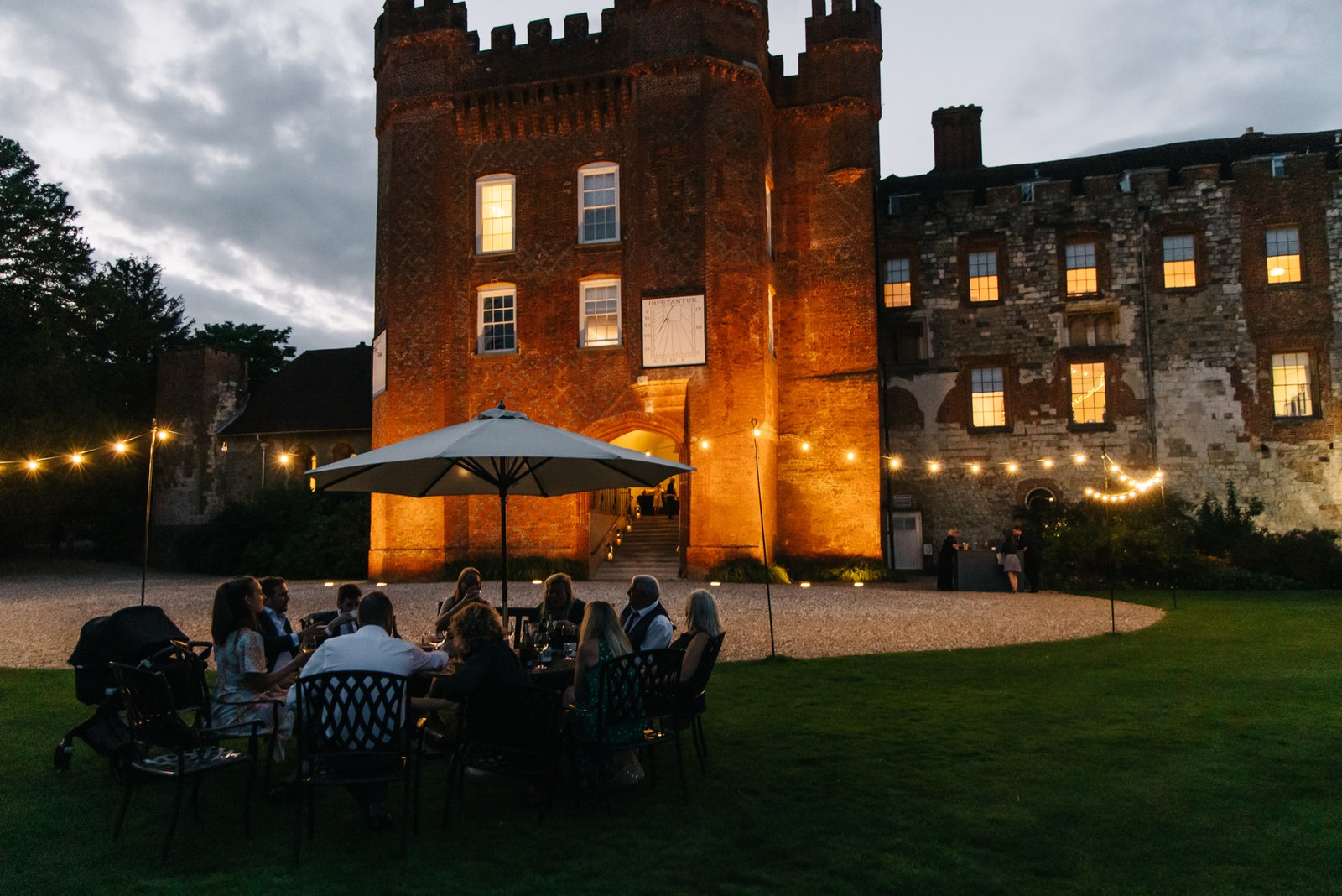 Farnham Castle floodlit at dusk, guests dining under a parasol on the lawn