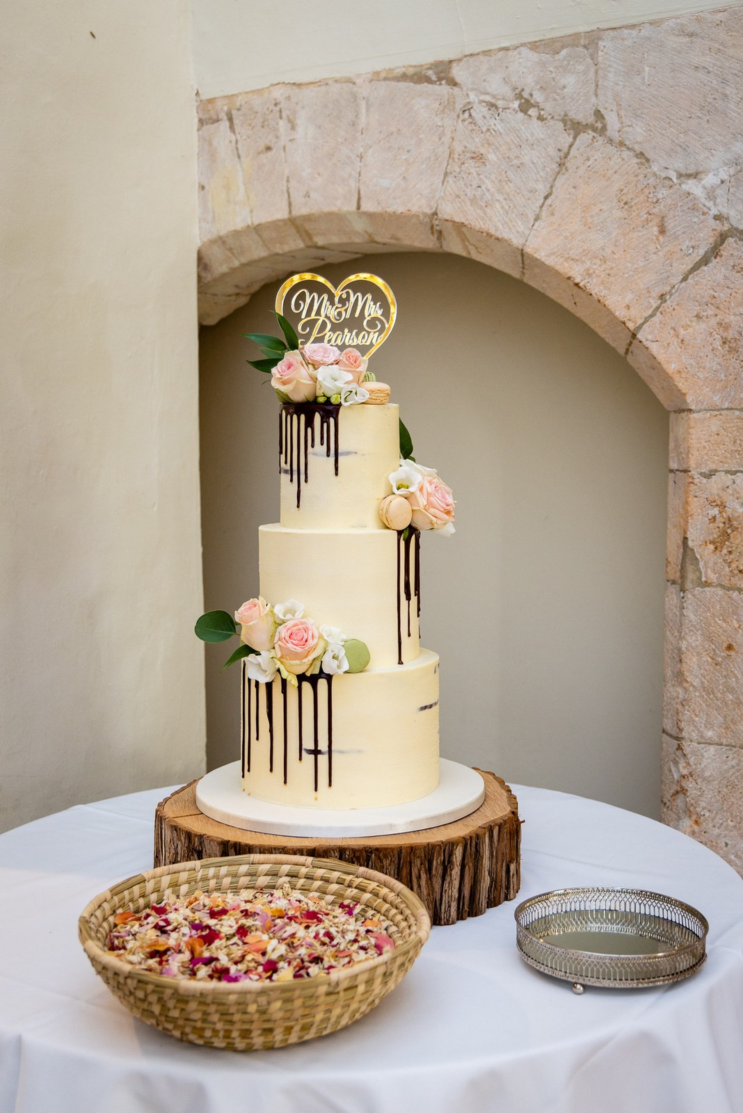 A tiered white cake with chocolate drip and roses, topped with a personalised sign, in a stone alcove