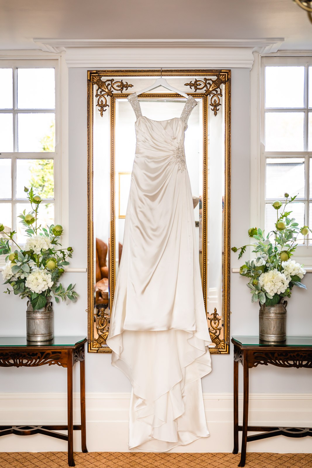 The satin wedding gown hanging on a gilt mirror between two floral urns at Fennes