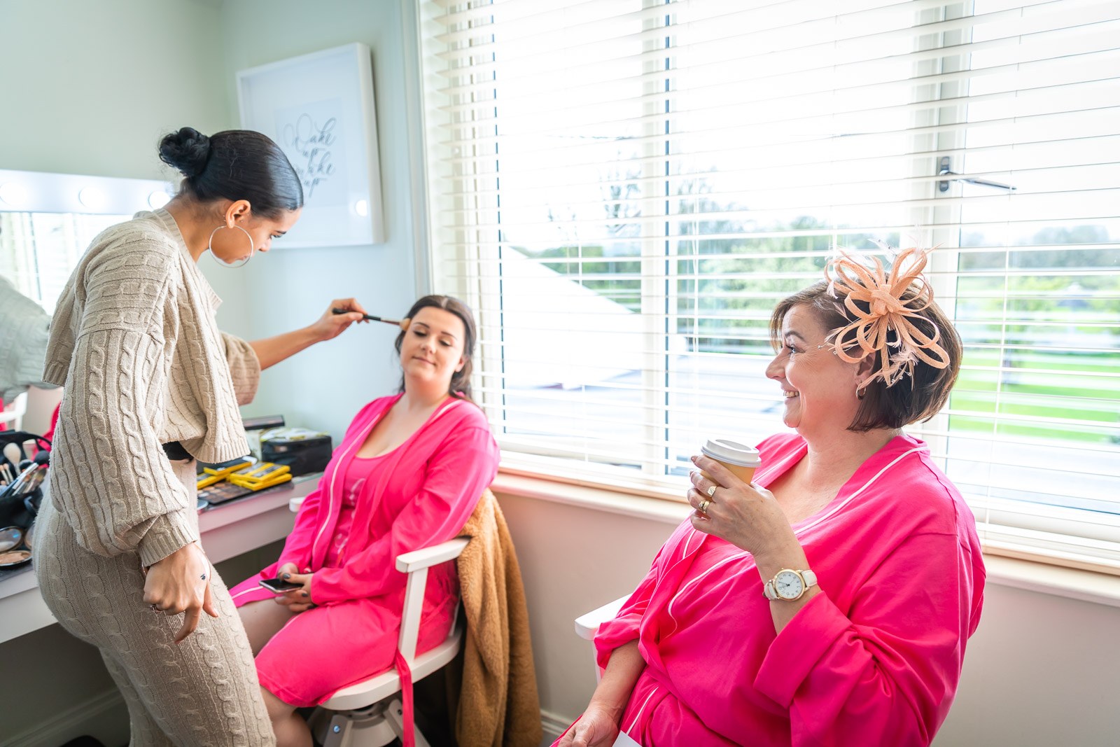 Makeup artist working on a bridesmaid as another looks on in a pink robe